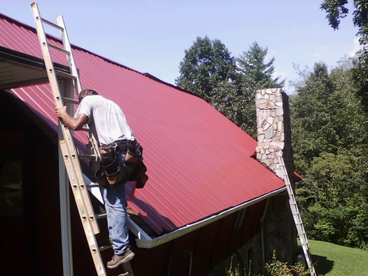 Roofer on a ladder beside a red metal roof and stone chimney — Soffit & Fascia Repair by Roofing Brothers in Thompson Falls